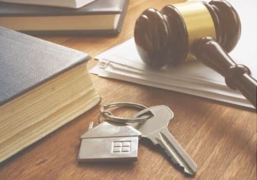 A house key with a little silver house lays on a wooden table next to an old book and a law gavel on top of a stack of papers.