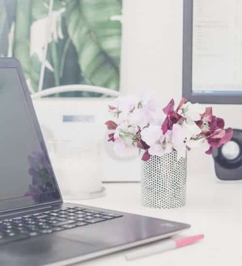 A laptop computer on a white desk with a small vase of pink flowers. Green picture on wall behind.